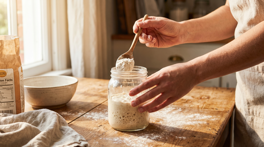 Hands feeding a sourdough starter in a glass jar with flour